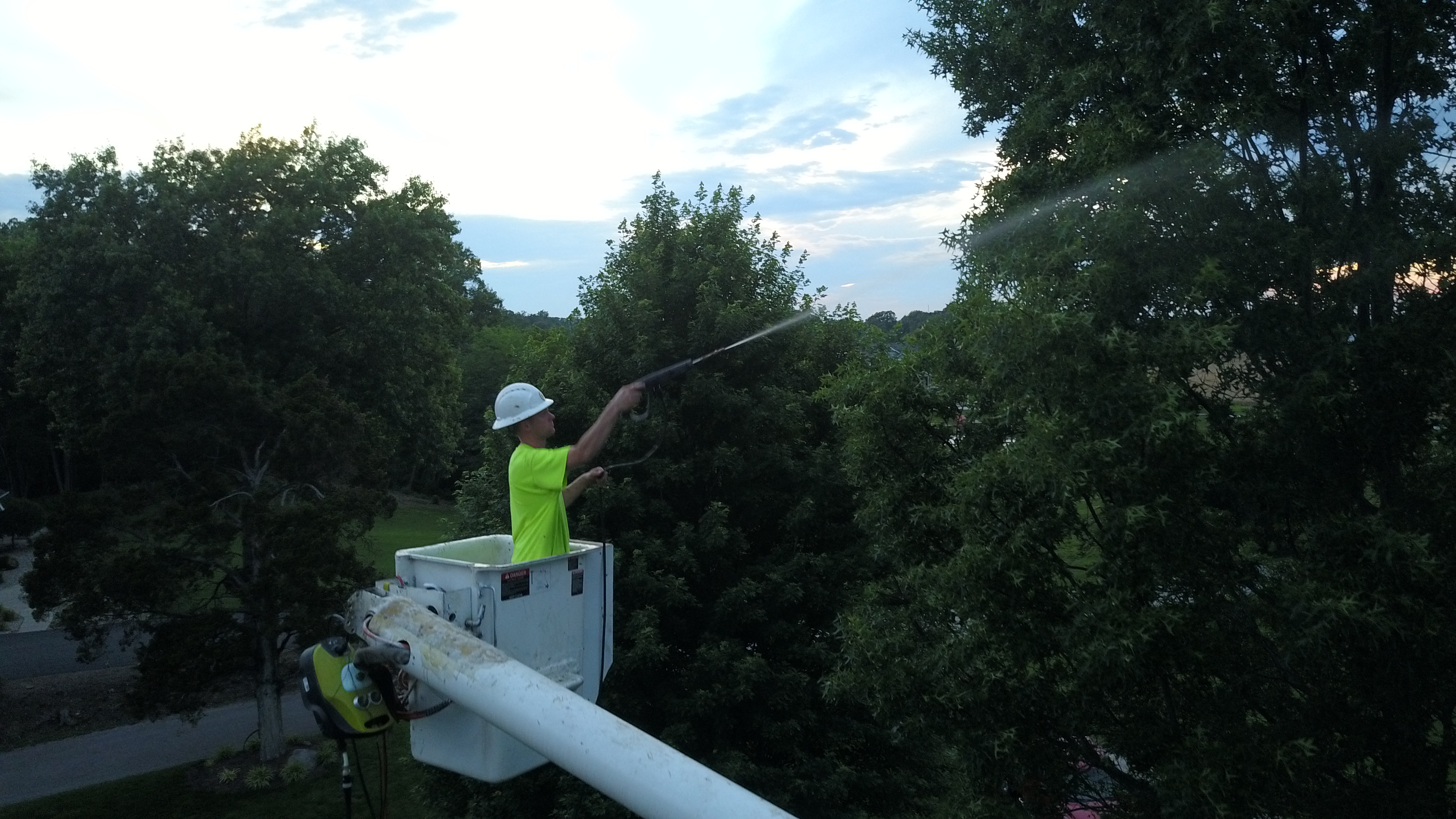 Close-up drone shot of Only Trees crew member in yellow vest and hard hat spraying tree treatment from bucket truck boom at dusk